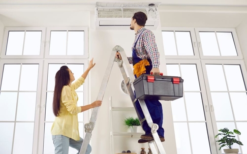 Woman pointing to AC while repairman stands on a latter